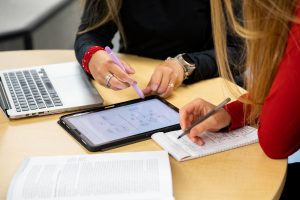 Two individuals at a table use pens and notepads, ipad, and laptop.